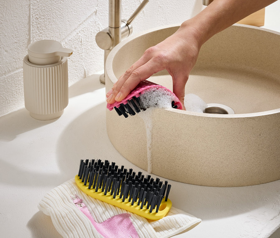 Une main nettoie un lavabo beige avec une brosse rose. Une brosse jaune et un tissu avec un flamant rose sont à côté.