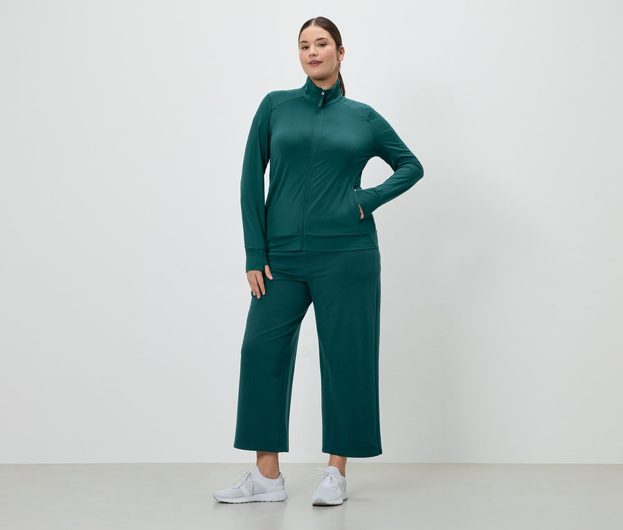 Femme portant un survêtement vert émeraude avec une veste zippée et une culotte, ainsi que des baskets blanches.