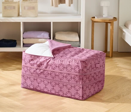 Sur un plancher en bois, un sac de rangement rose à motif géométrique, partiellement rempli d'une couette blanche.