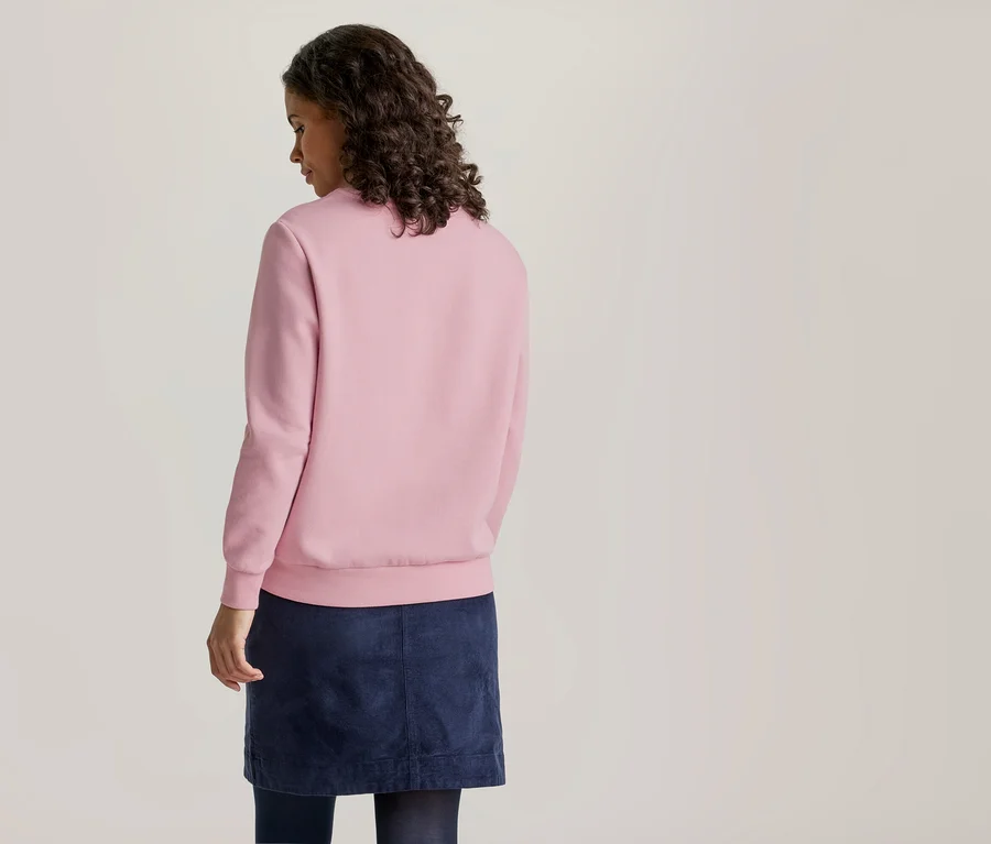 Une femme aux cheveux foncés et bouclés se tient dos au spectateur, regardant légèrement vers la droite. Elle porte un sweat-shirt rose uni à manches longues et une jupe bleu marine.