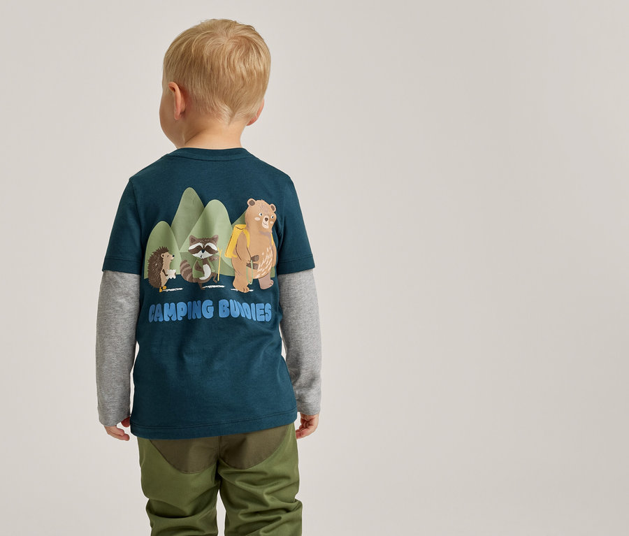 Un jeune enfant, vu de dos, porte un t-shirt bleu sarcelle foncé sur un t-shirt gris à manches longues et un pantalon vert olive. Le t-shirt présente un graphique d'un hérisson, d'un raton laveur et d'un ours devant des montagnes vertes, avec le texte 'CAMPING BUNNIES' en dessous.