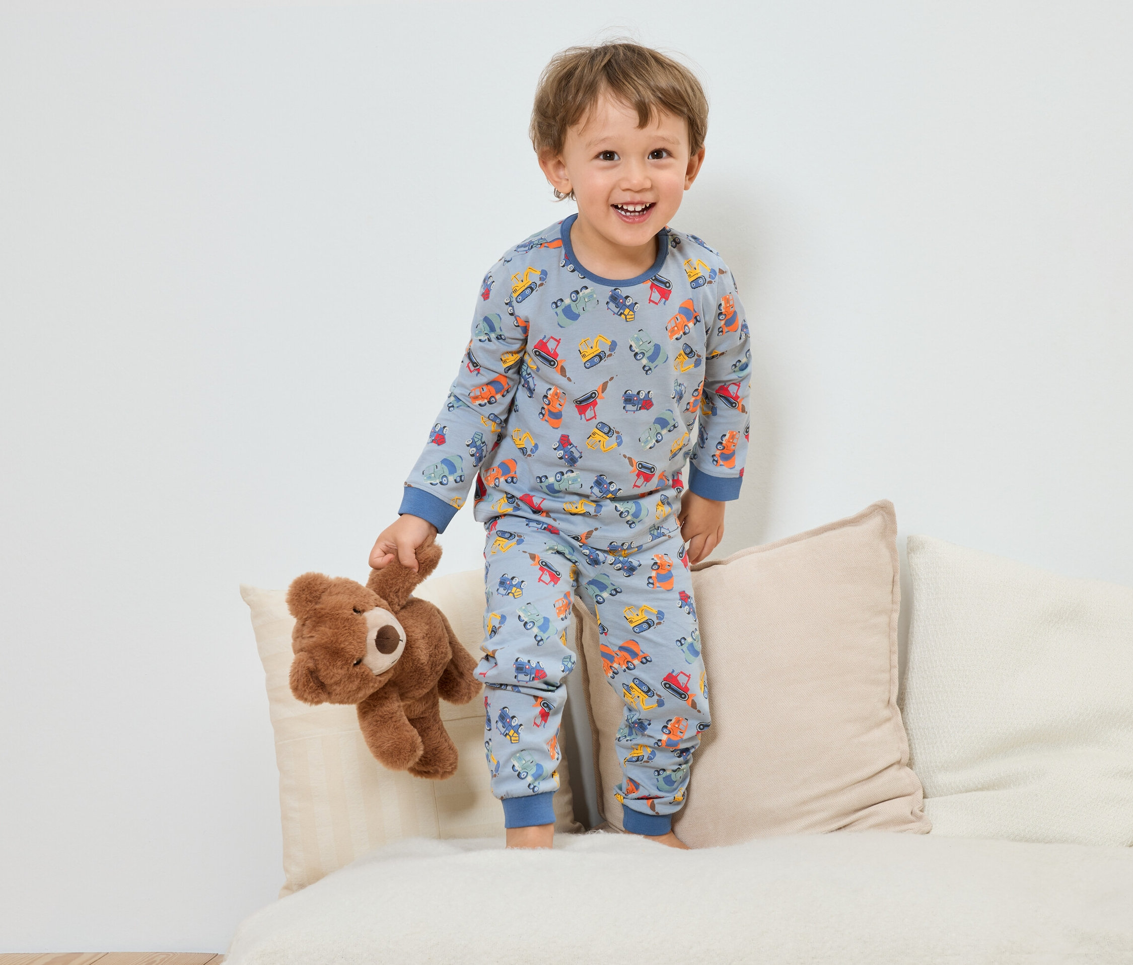 Un garçon souriant en pyjama bleu avec un motif de véhicules de construction se tient debout avec un ours en peluche sur un canapé.