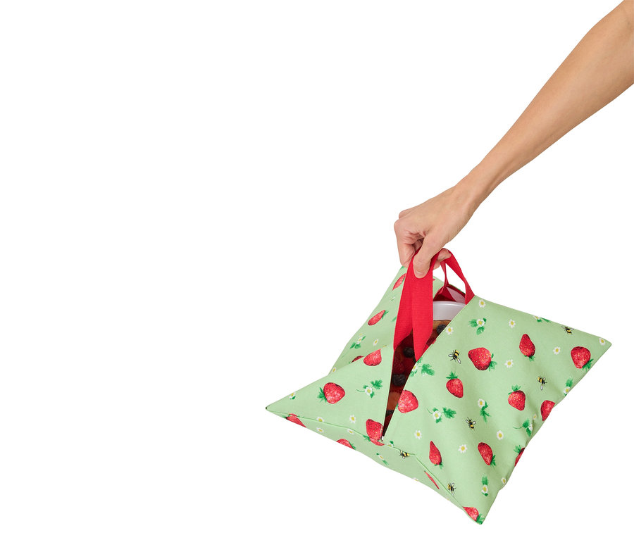 Une main tient un sac vert clair à motifs de fraises avec des anses rouges, contenant un récipient avec de la nourriture.