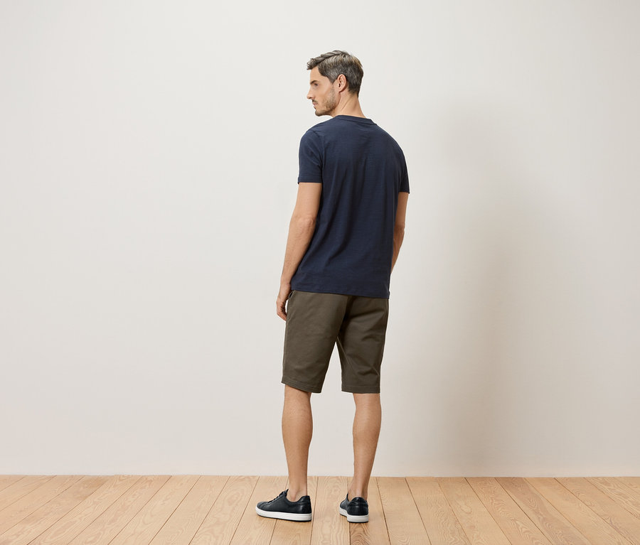 Un homme se tient dos à la caméra, portant un t-shirt bleu marine, un short kaki et des chaussures bleu marine.