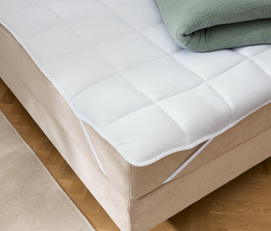 Un protège-matelas blanc matelassé avec des élastiques aux coins est posé sur un lit beige. Une couverture verte pliée est posée sur le dessus.