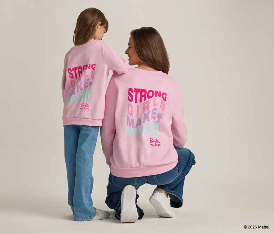 Vue arrière d'une jeune femme agenouillée à côté d'une jeune fille. Toutes deux portent des pulls rose clair avec l'inscription "Strong Girls Make Music".