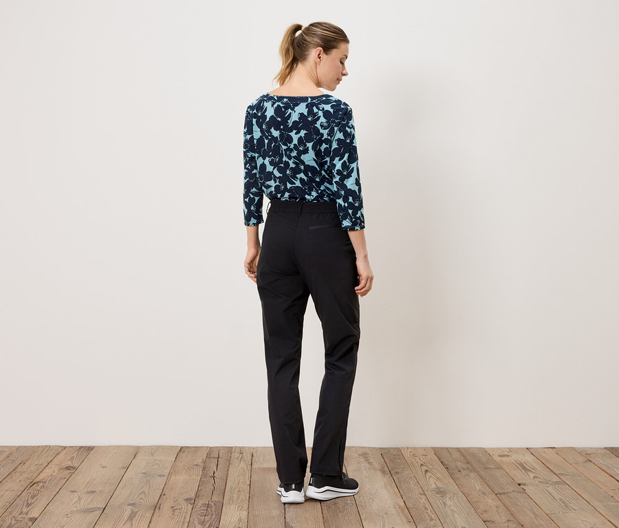 Une femme, dos à la caméra, porte un chemisier bleu marine à motifs floraux et un pantalon noir.