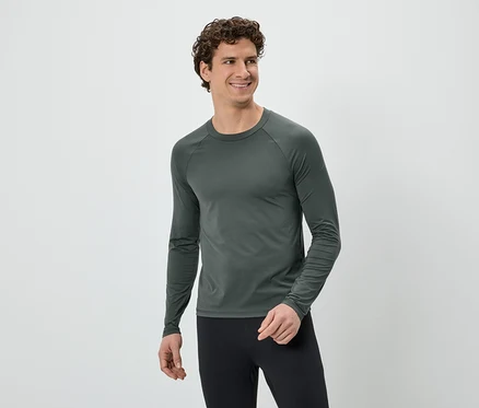 Un homme souriant aux cheveux bouclés, portant un t-shirt gris à manches longues et un legging noir, regarde vers la droite sur un fond blanc.