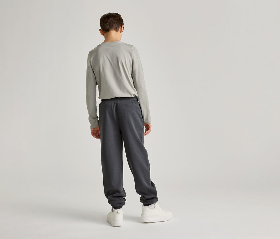 Un enfant portant un T-shirt à manches longues gris clair et un pantalon de jogging gris foncé, dos au spectateur, sur un fond clair.