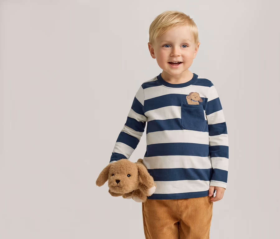Un garçon souriant aux cheveux blonds tient un chien en peluche. Il porte un t-shirt à manches longues rayé bleu et blanc avec une broderie de chien sur la poche.