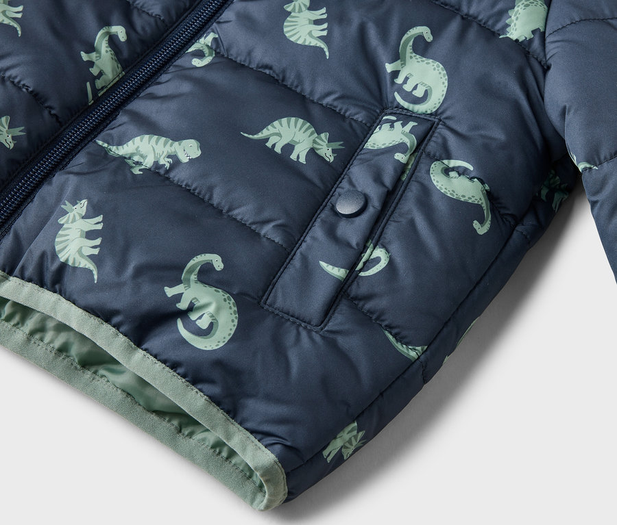 Gros plan d'une veste matelassée bleu marine avec un motif de dinosaures vert clair, une fermeture éclair et une poche à bouton-pression.