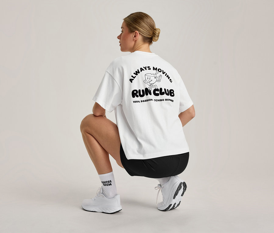 Une femme aux cheveux blonds attachés en chignon est accroupie, vue de dos. Elle porte un T-shirt de sport blanc oversize avec un motif noir 'ALWAYS MOVING RUN CLUB' dans le dos, un short noir, des chaussettes blanches et des chaussures de sport blanches.