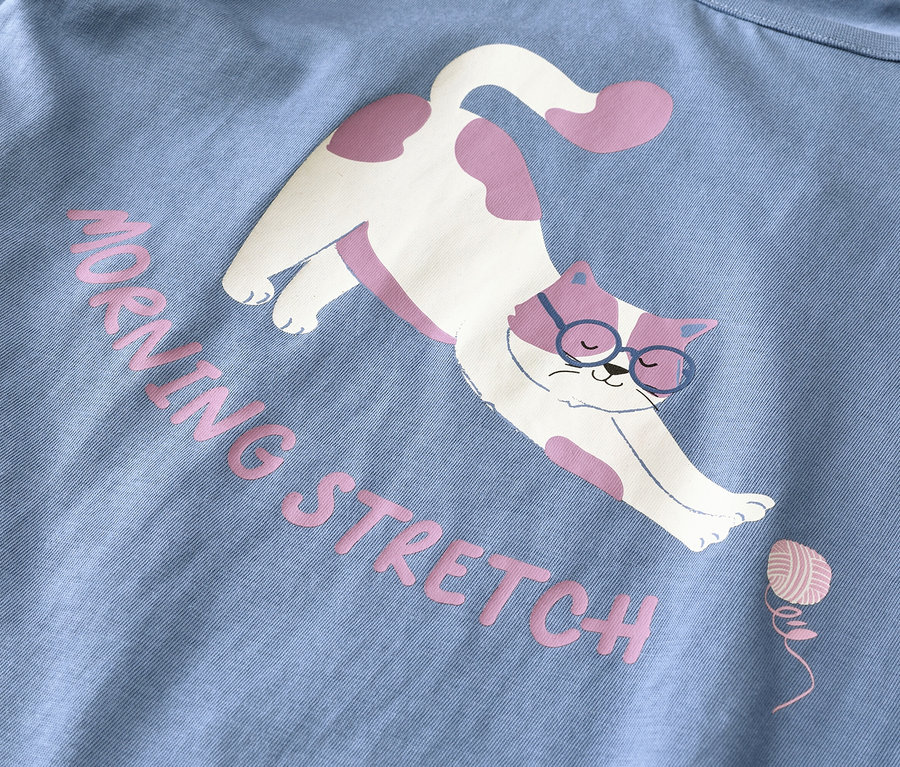 Tissu bleu clair avec un motif d'un chat blanc à taches roses portant des lunettes bleues, s'étirant. Le texte rose indique « MORNING STRETCH » à côté d'une petite pelote de laine rose.