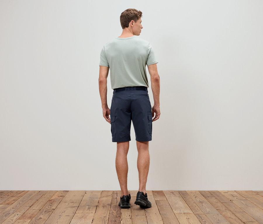 Un homme vu de dos, portant un t-shirt vert olive et un short cargo bleu, se tient sur un plancher en bois devant un mur blanc.