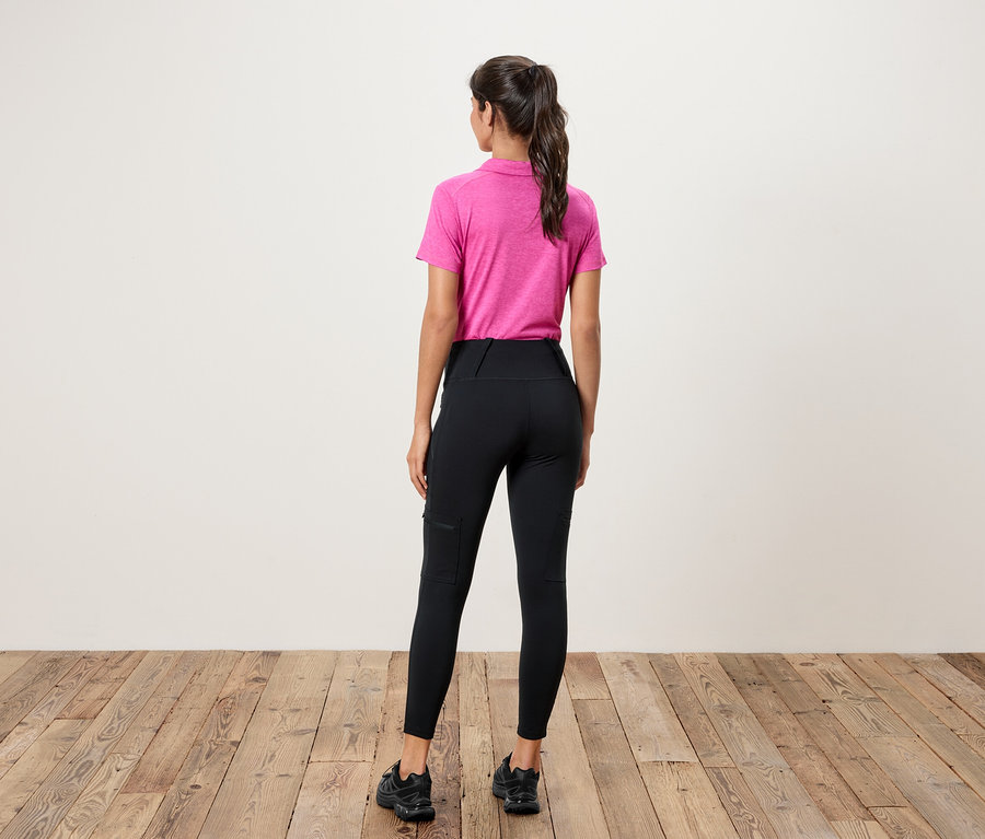 Une femme se tient dos à la caméra, portant un polo rose et un legging noir avec des poches.