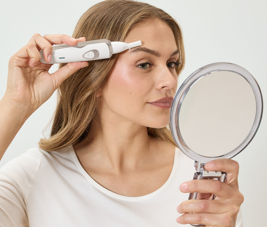 Une femme s'épile les sourcils avec un épilateur blanc tout en regardant dans un miroir rond.