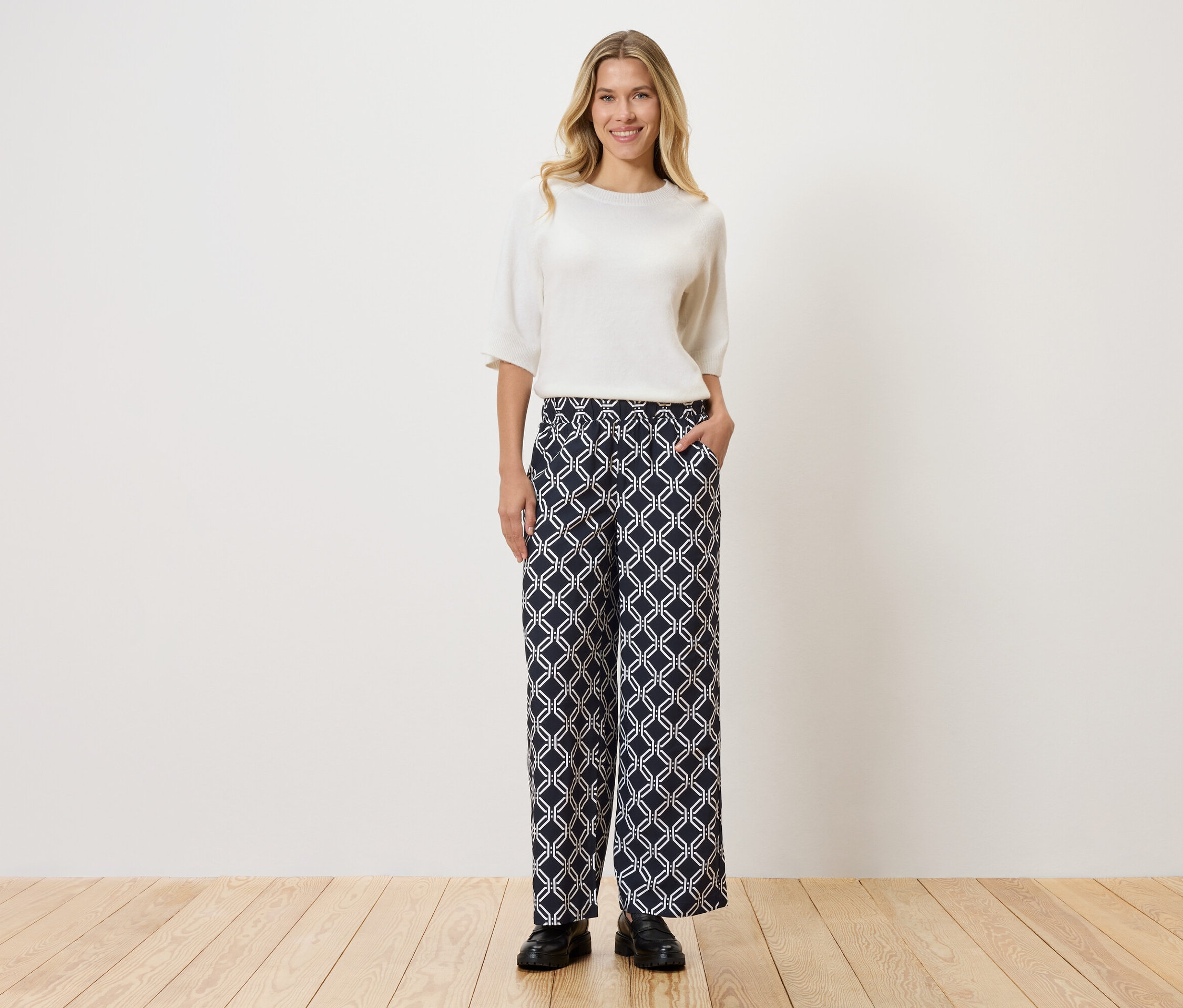 Femme portant un haut blanc et un pantalon à motifs, debout sur un plancher en bois devant un mur.