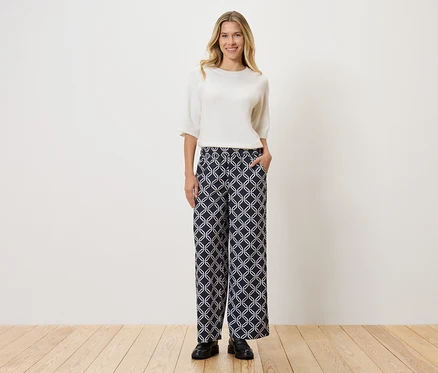 Femme portant un haut blanc et un pantalon à motifs, debout sur un plancher en bois devant un mur.