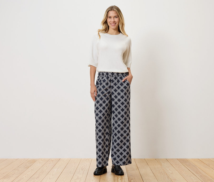 Femme portant un haut blanc et un pantalon à motifs, debout sur un plancher en bois devant un mur.