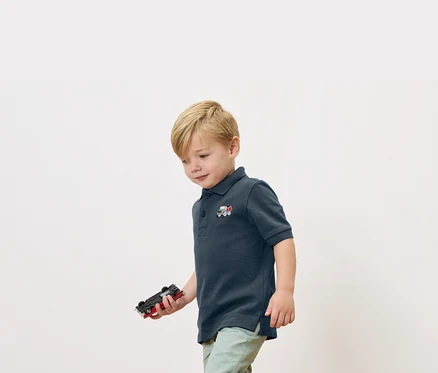 Un garçon blond tient une voiture jouet à la main et porte un polo bleu marine avec une petite application de train.