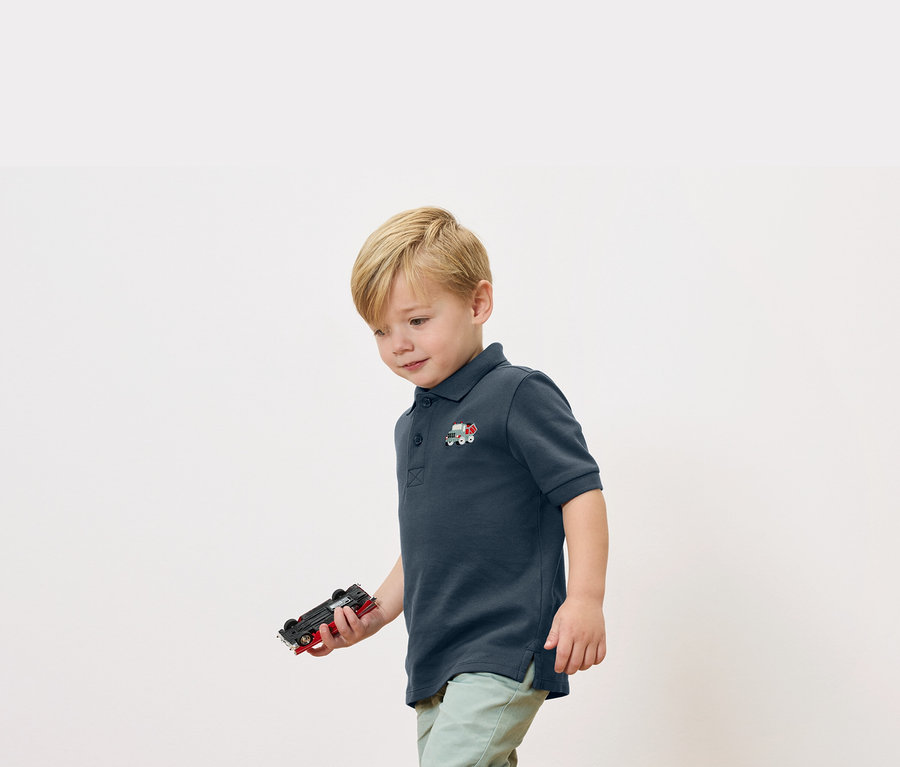 Un garçon blond tient une voiture jouet à la main et porte un polo bleu marine avec une petite application de train.