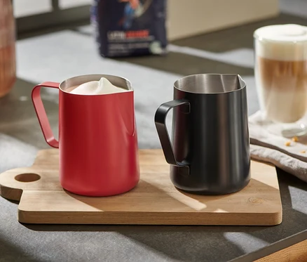 Un pichet à lait rouge avec du lait mousseux et un pichet à lait noir sur une planche à bois.