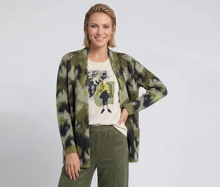 Femme souriante aux cheveux blonds courts portant un cardigan vert à motifs, un t-shirt clair avec un imprimé graphique et un pantalon vert olive.
