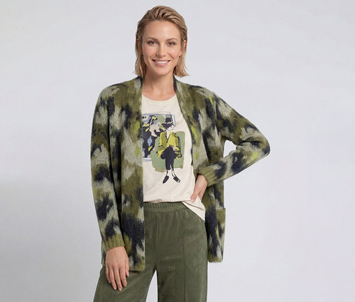 Femme souriante aux cheveux blonds courts portant un cardigan vert à motifs, un t-shirt clair avec un imprimé graphique et un pantalon vert olive.