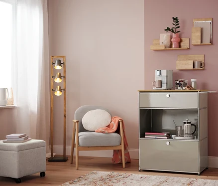 Salon avec fauteuil, lampe et commode avec machine à café. Sur la commode, un panier à capsules, blanc.