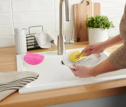 Une femme lave une assiette avec une éponge en silicone jaune dans un évier blanc. Une éponge rose est à côté.