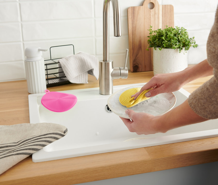Une femme lave une assiette avec une éponge en silicone jaune dans un évier blanc. Une éponge rose est à côté.