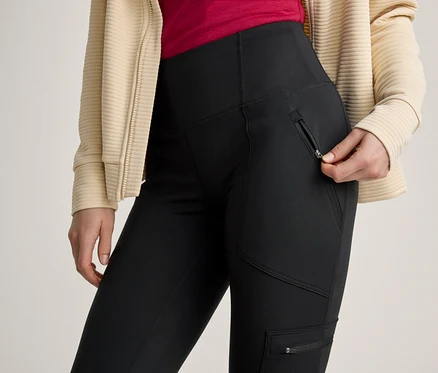 Une personne vêtue d'un pantalon noir à poches zippées et d'une veste côtelée beige ouvre la fermeture éclair d'une poche latérale du pantalon.