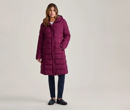 Femme debout, portant un long manteau matelassé violet à capuche, un jean foncé et des mocassins noirs, les mains dans les poches.