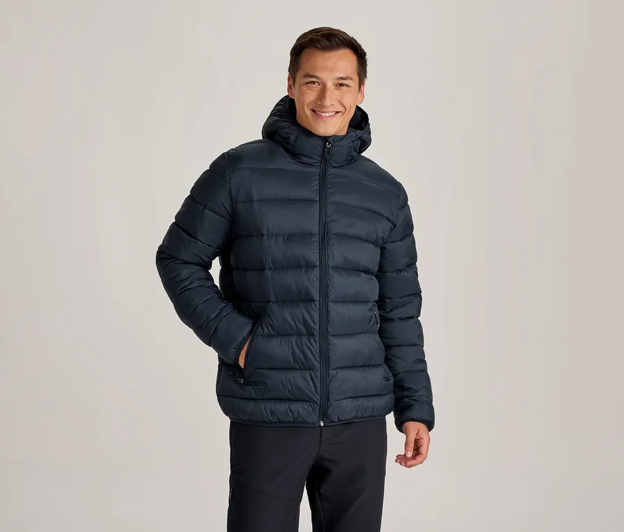 Homme souriant portant une doudoune à capuche bleu foncé et un pantalon noir, une main dans la poche de sa veste.