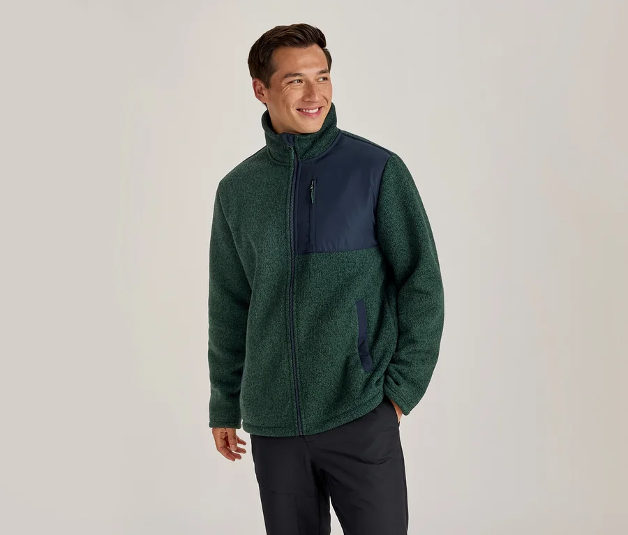 Homme portant une veste polaire verte et bleu marine et un pantalon noir, souriant et regardant vers la droite.