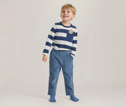 Jeune garçon debout, regardant vers le haut et souriant, portant un t-shirt rayé bleu marine et blanc à manches longues avec une poche poitrine à motif animal, un pantalon en velours côtelé bleu et des chaussettes bleues.