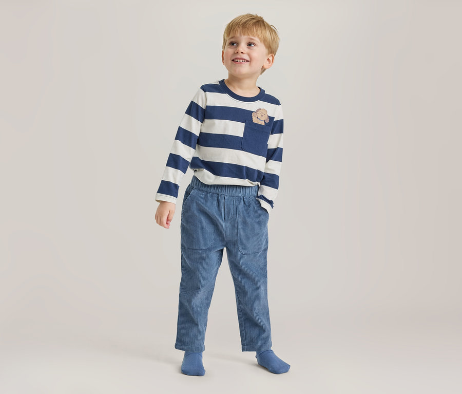 Jeune garçon debout, regardant vers le haut et souriant, portant un t-shirt rayé bleu marine et blanc à manches longues avec une poche poitrine à motif animal, un pantalon en velours côtelé bleu et des chaussettes bleues.