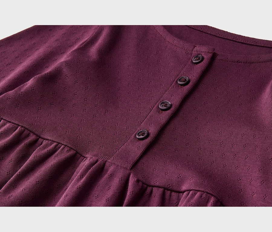Gros plan d'un vêtement en tricot bordeaux avec un motif texturé subtil. Il présente un col rond, une patte de boutonnage avec quatre boutons foncés et une couture froncée sous la poitrine.