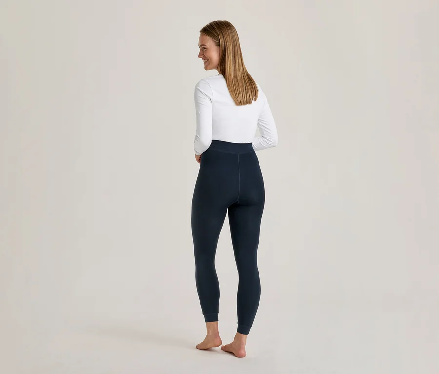 Femme de dos, souriant et regardant par-dessus son épaule, vêtue d'un haut blanc à manches longues et d'un legging bleu foncé.