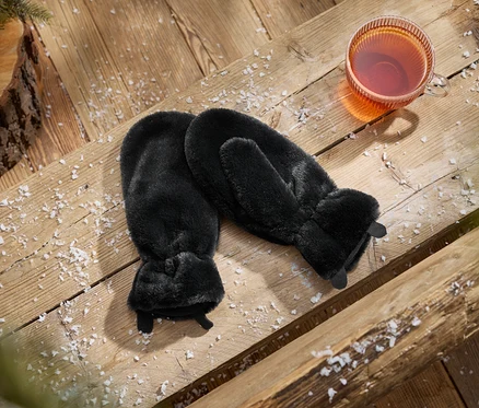 Une paire de moufles noires en fausse fourrure à côté d'une tasse de thé sur une surface en bois saupoudrée de neige, avec une pomme de pin et une branche.