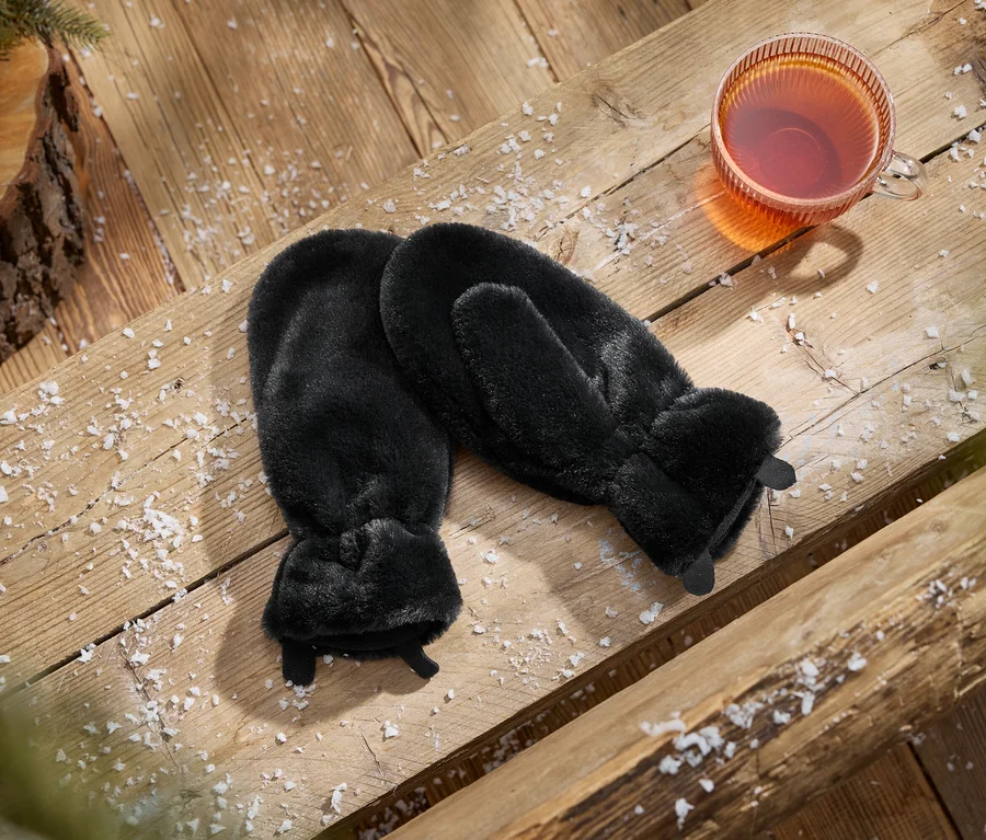 Une paire de moufles noires en fausse fourrure à côté d'une tasse de thé sur une surface en bois saupoudrée de neige, avec une pomme de pin et une branche.