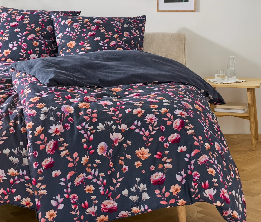 Lit avec une parure de lit bleu foncé à motifs floraux roses, oranges et blancs. Une table de chevet en bois clair avec une bouteille d'eau et un verre est à droite. Un cadre et une suspension rose sont au-dessus.