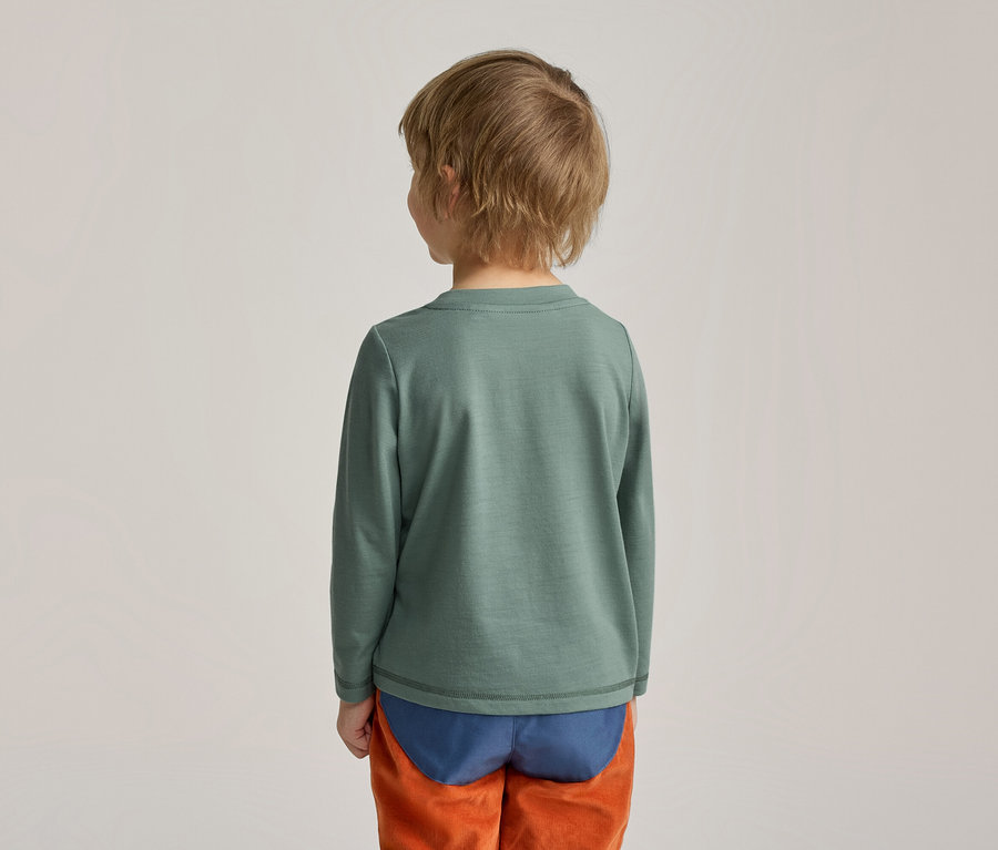 Un enfant vu de dos, portant un t-shirt vert à manches longues, des sous-vêtements bleus et un pantalon orange.