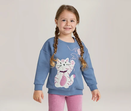 Jeune fille souriante avec des tresses, portant un sweat-shirt bleu avec une application de chat blanc et rose et un pantalon côtelé rose.