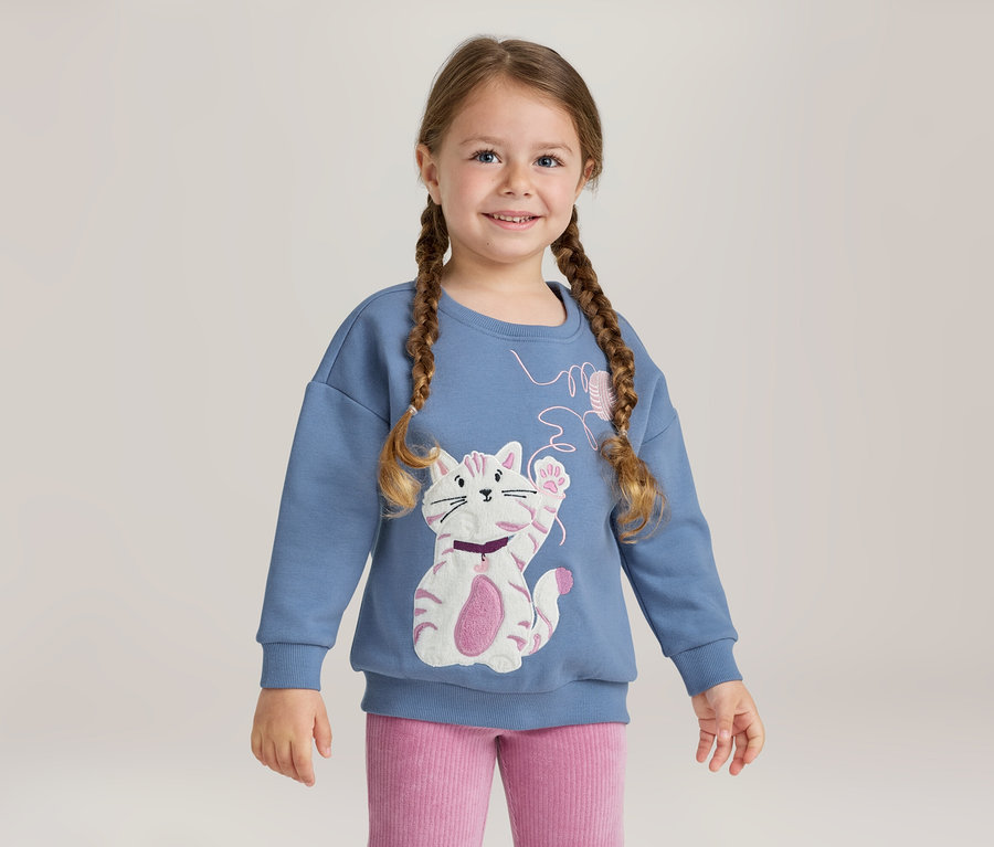 Jeune fille souriante avec des tresses, portant un sweat-shirt bleu avec une application de chat blanc et rose et un pantalon côtelé rose.