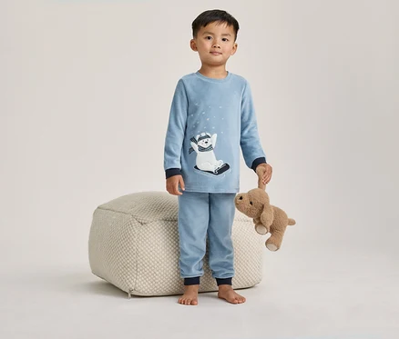 Jeune garçon en pyjama bleu avec un motif d'ours polaire, tenant un chien en peluche marron, debout à côté d'un pouf clair.