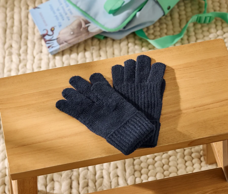 Des gants tricotés bleu foncé sont posés sur un tabouret en bois clair. À l'arrière-plan, un tapis en grosse maille crème, un sac bleu clair et un livre illustré d'un éléphant sont visibles.