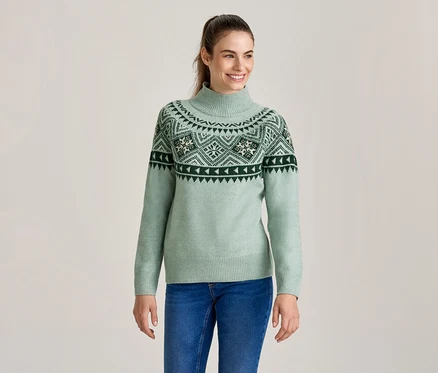 Femme souriante portant un pull à col roulé vert clair avec un motif Fair Isle vert foncé et blanc, et un jean bleu.