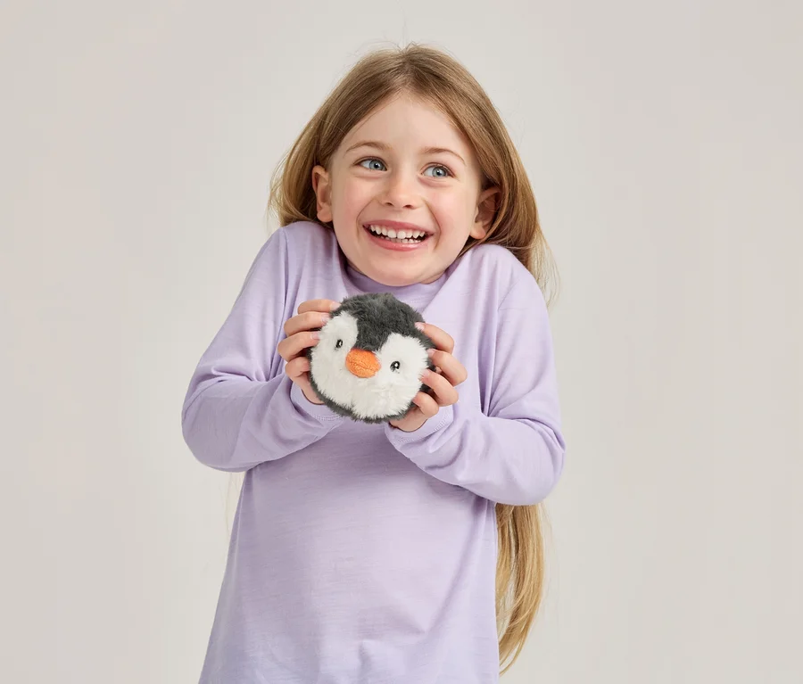 Une jeune fille souriante aux longs cheveux blonds et aux yeux bleus, vêtue d'un t-shirt violet clair à manches longues, tient un jouet pingouin rond en peluche.