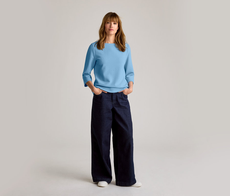 Femme aux cheveux mi-longs, portant un haut bleu clair, un jean large bleu foncé et des baskets en cuir blanches.